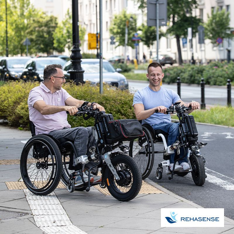 Zwei Männer im Rollstuhl benutzen das elektrische Zugsystem PAWS von Rehasense und fahren damit in der Stadt.