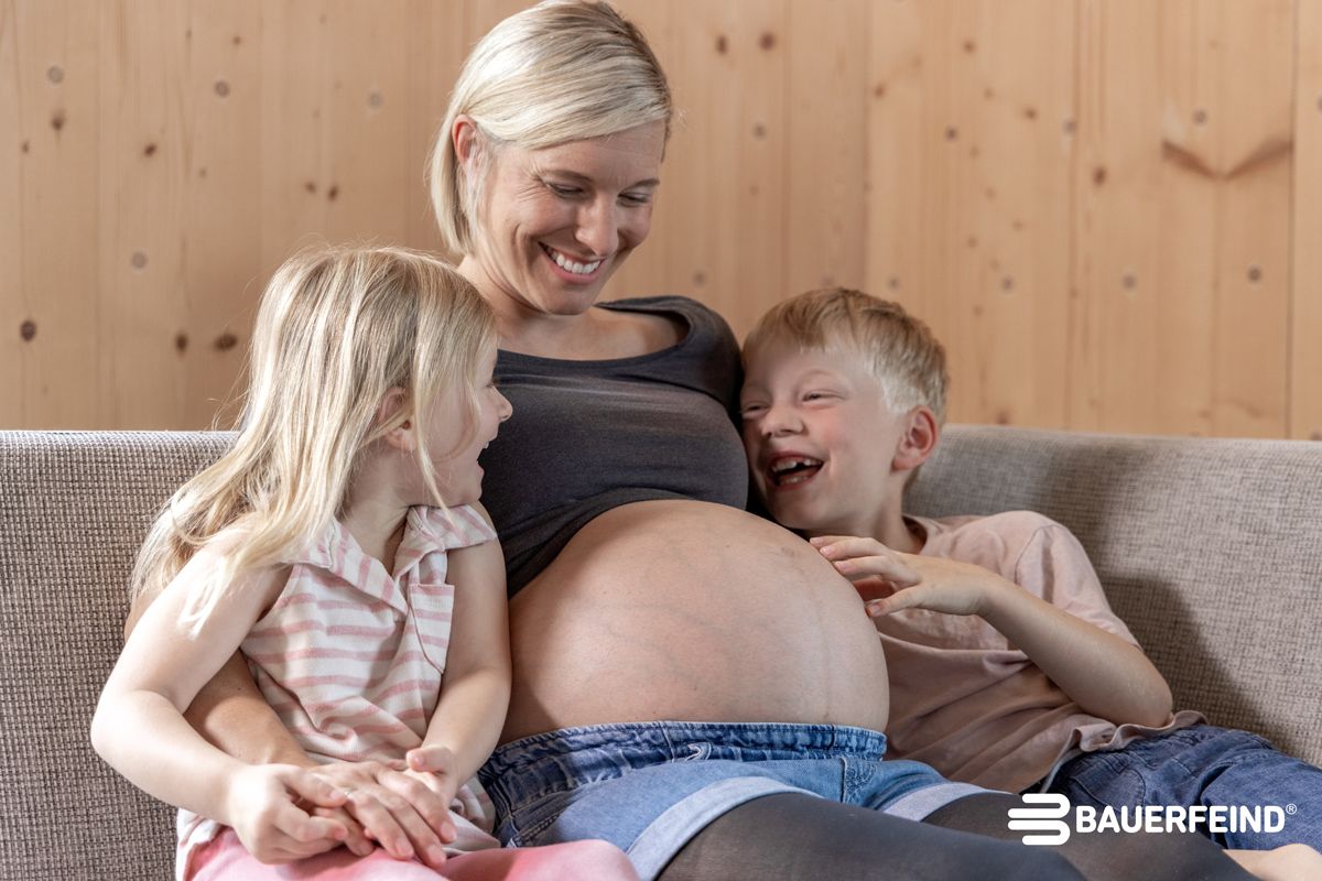 Eine schwangere Frau sitzt mit ihren zwei Kindern auf dem Sofa und trägt eine Strumpfhose zur Kompression in der Schwangerschaft von Bauerfeind.