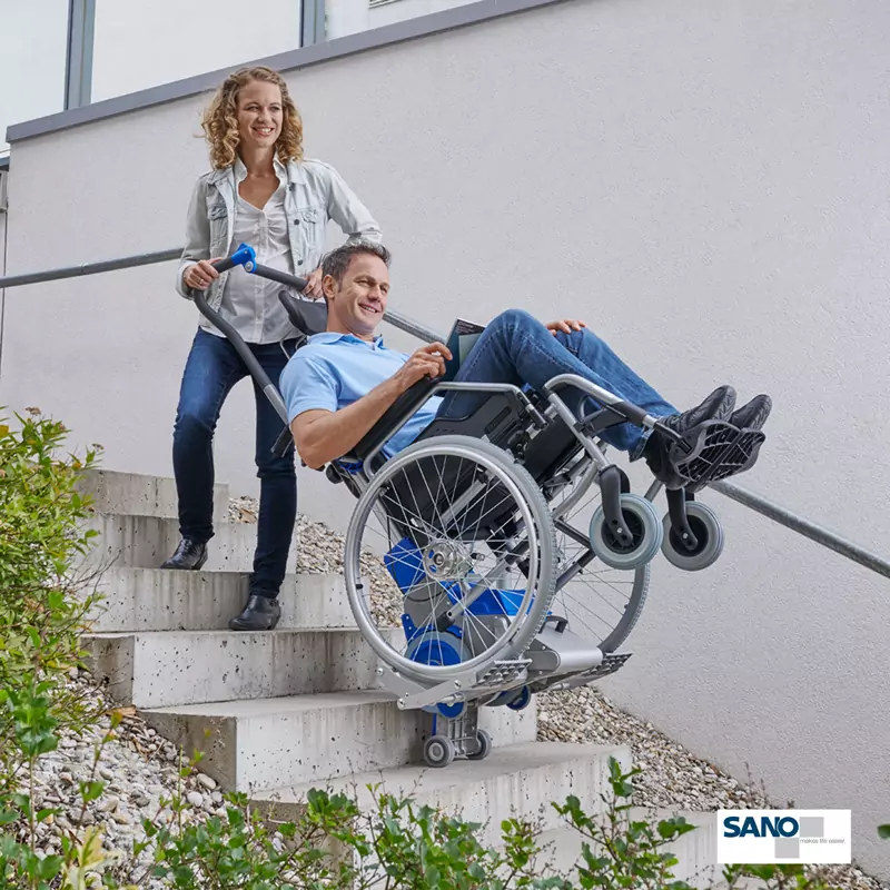 Eine Frau zieht einen im Rollstuhl sitzenden Mann mithilfe eines Rechatechnikhilfsmittels den Liftkar die Treppe hoch.