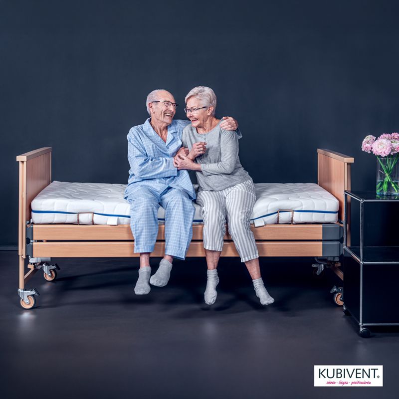 Auf einem Pflegebett von Kubivent sitzt ein Seniorenpaar, das sich umarmt und lacht. Hilfsmittel für die Reha-Technik.