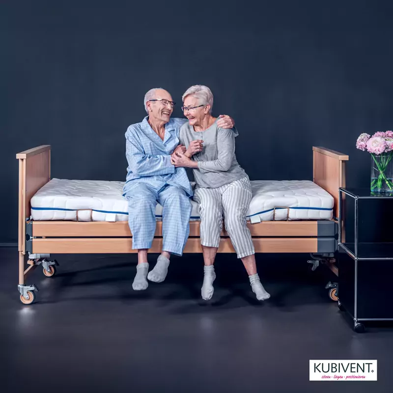 Auf einem Pflegebett von Kubivent sitzt ein Seniorenpaar, das sich umarmt und lacht. Hilfsmittel für die Reha-Technik.