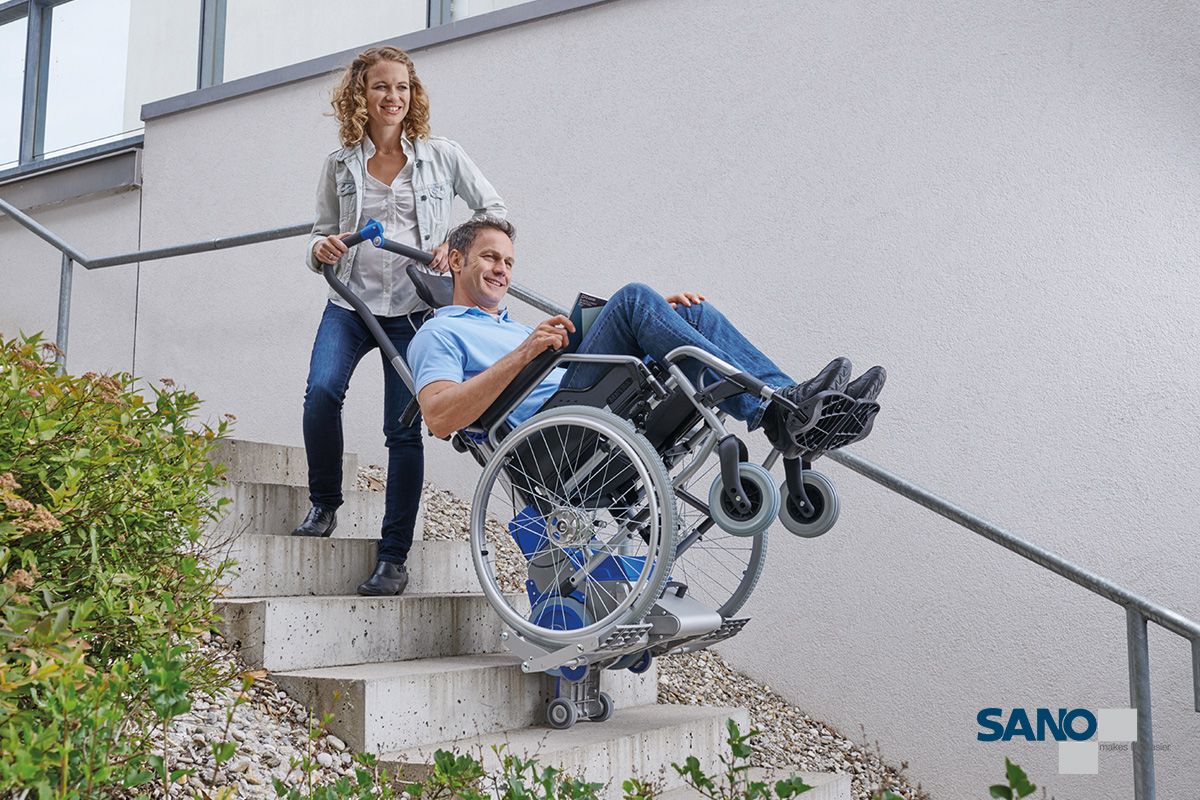Eine Frau zieht einen im Rollstuhl sitzenden Mann mithilfe eines Treppensteiger den Liftkar PT von Sano die Treppe hoch.