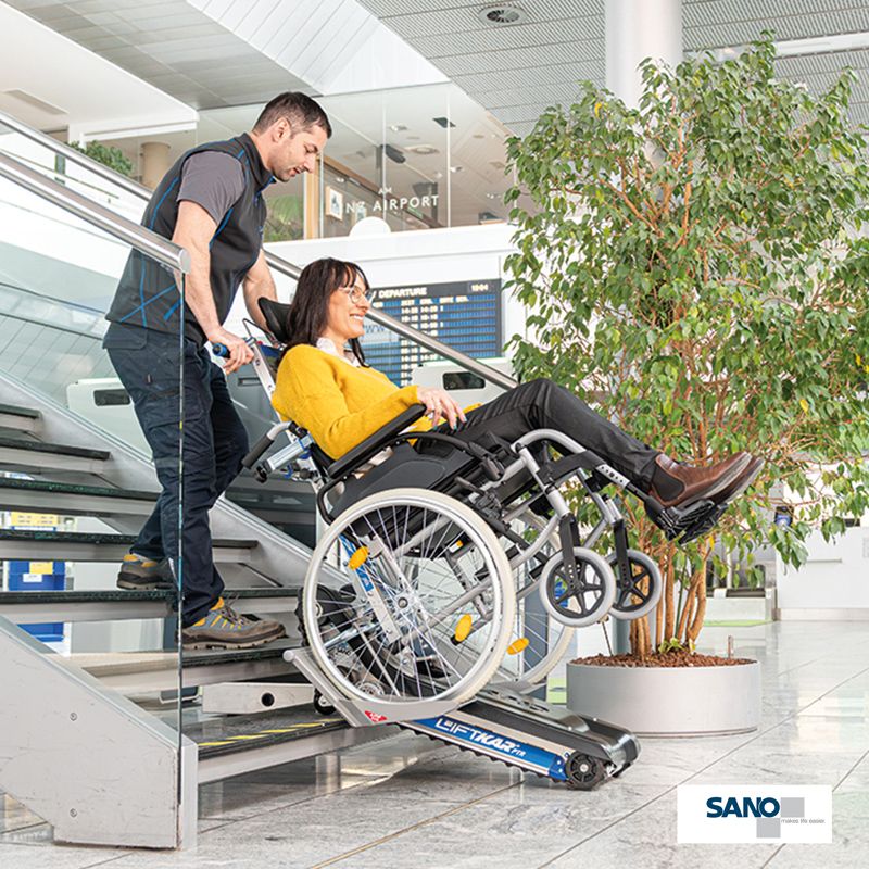 Ein Mann zieht eine im Rollstuhl sitzende Frau mithilfe eines Treppenraupen den Liftkar PTR von Sano die Treppe hoch.