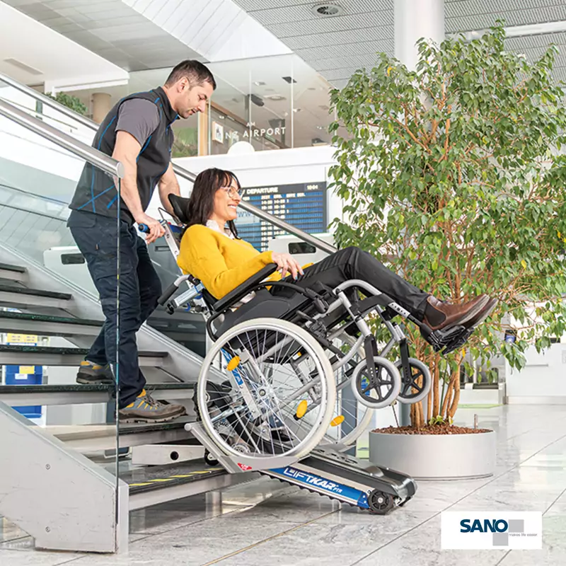 Ein Mann zieht eine im Rollstuhl sitzende Frau mithilfe eines Treppenraupen den Liftkar PTR von Sano die Treppe hoch.