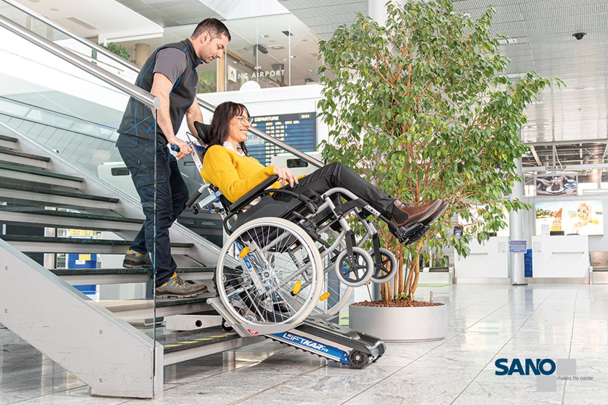 Ein Mann zieht eine im Rollstuhl sitzende Frau mithilfe eines Treppenraupen den Liftkar PTR von Sano die Treppe hoch.
