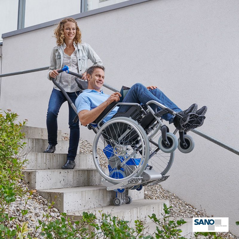 Eine Frau zieht einen im Rollstuhl sitzenden Mann mithilfe eines Rechatechnikhilfsmittels den Liftkar die Treppe hoch.