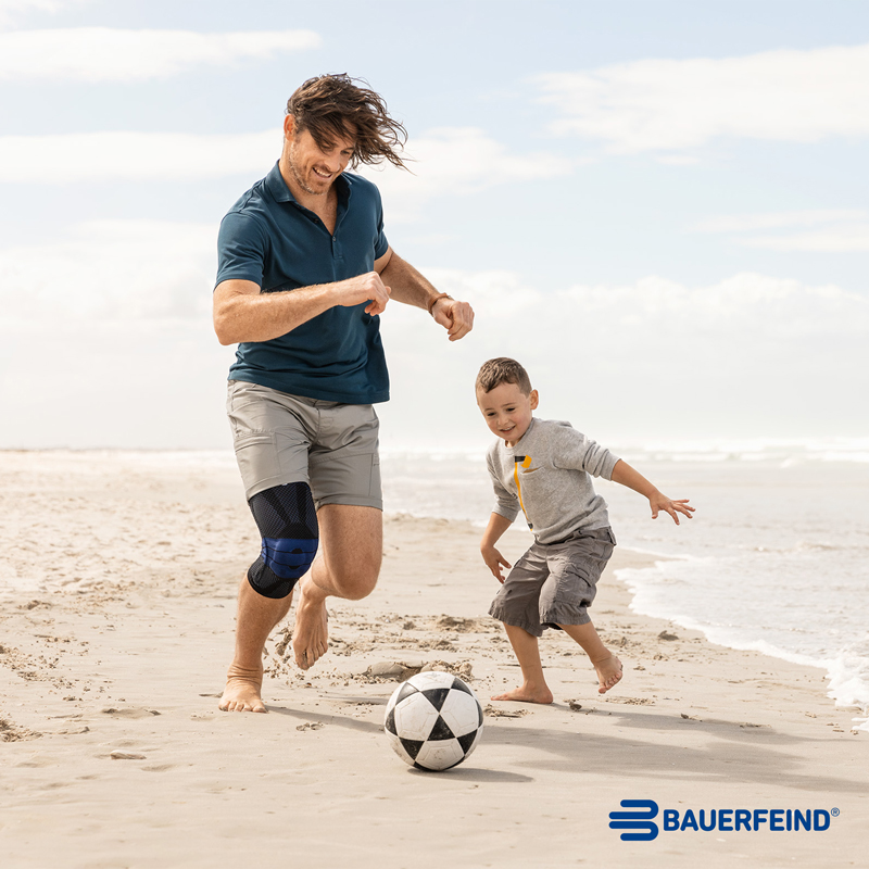 Ein Mann spielt mit seinem Sohn Fußball am Strand und trägt dabei die Kniebandage von Bauerfeind, die es in unserem Sanitätshandel in Nürnberg und Umgebung gibt.