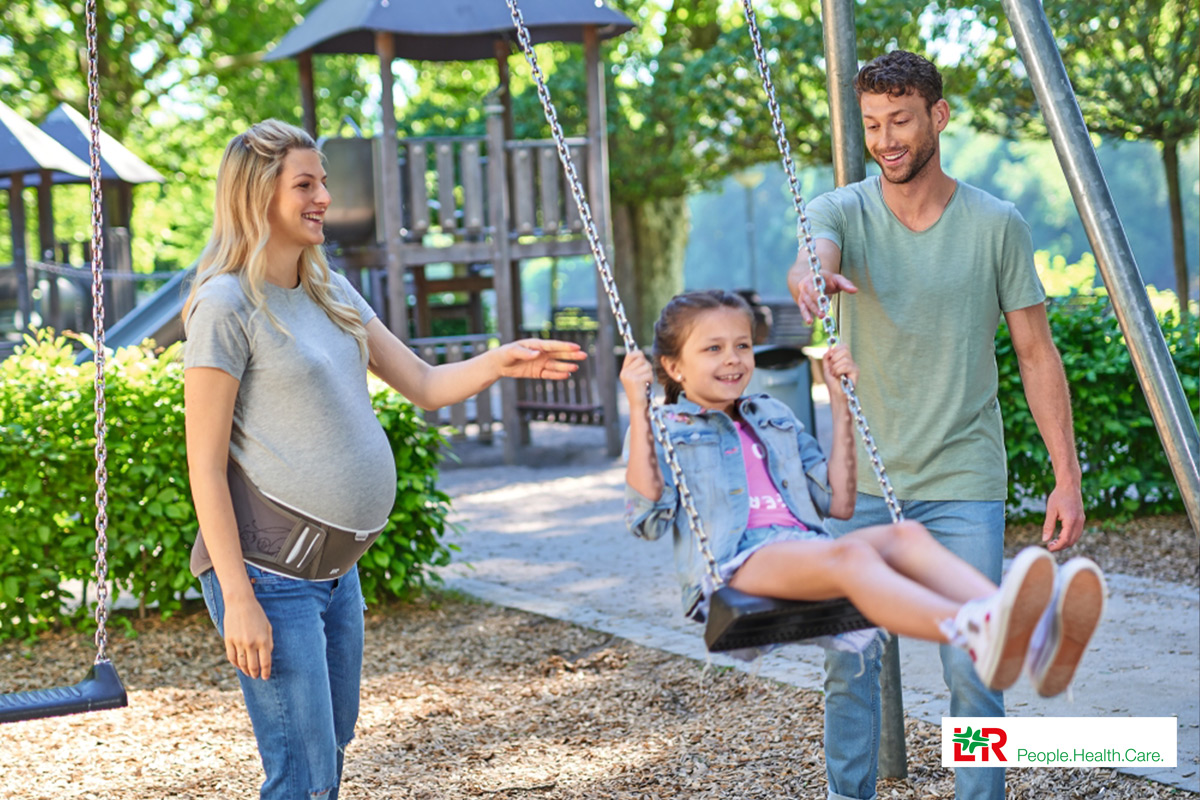 Materna Comfort Schwangerschafts-Rückenorthese Lohmann & Rauscher Eine schwangere Frau trägt eine Rückenorthese von L&R und spielt mit ihrem Kind und ihrem Mann auf dem Spielplatz.
