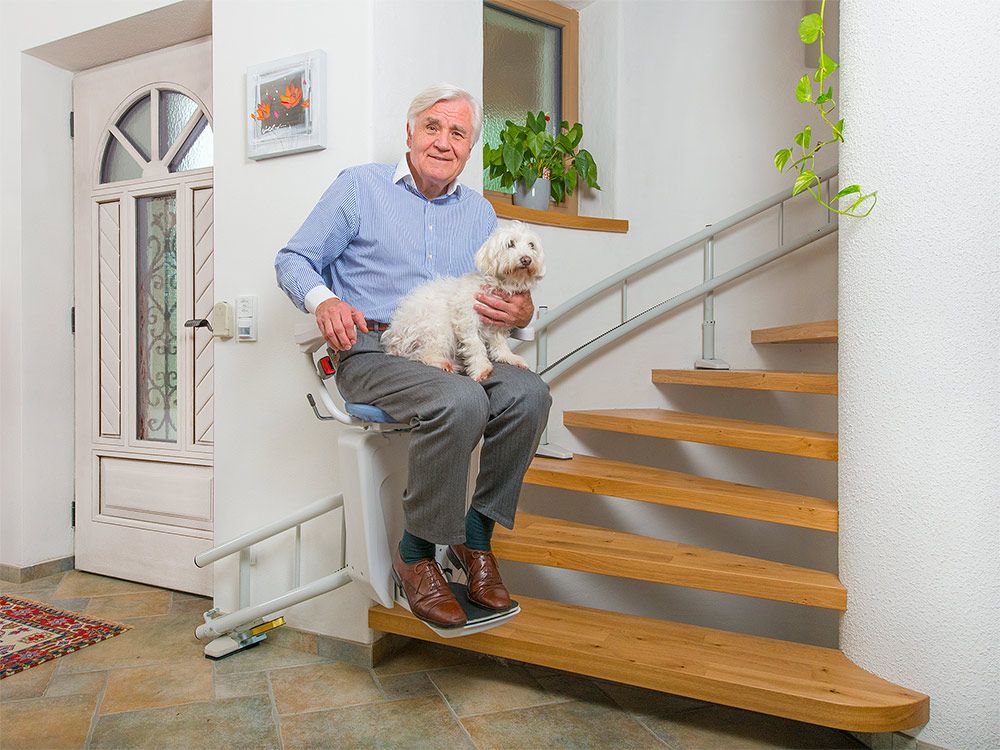 Ein Mann mit einem kleinem Hund auf dem Schoß benutzt den Treppenlift Lehner Alpha